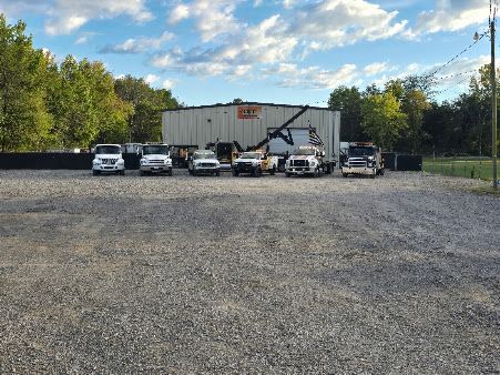 White utility building with parked trucks and an excavator on a gravel lot, surrounded by trees under a cloudy sky.