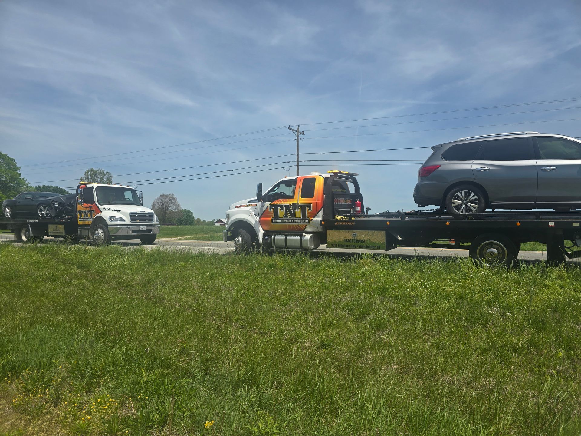 Tow truck hauling a silver SUV in a grassy roadside field under a blue sky.