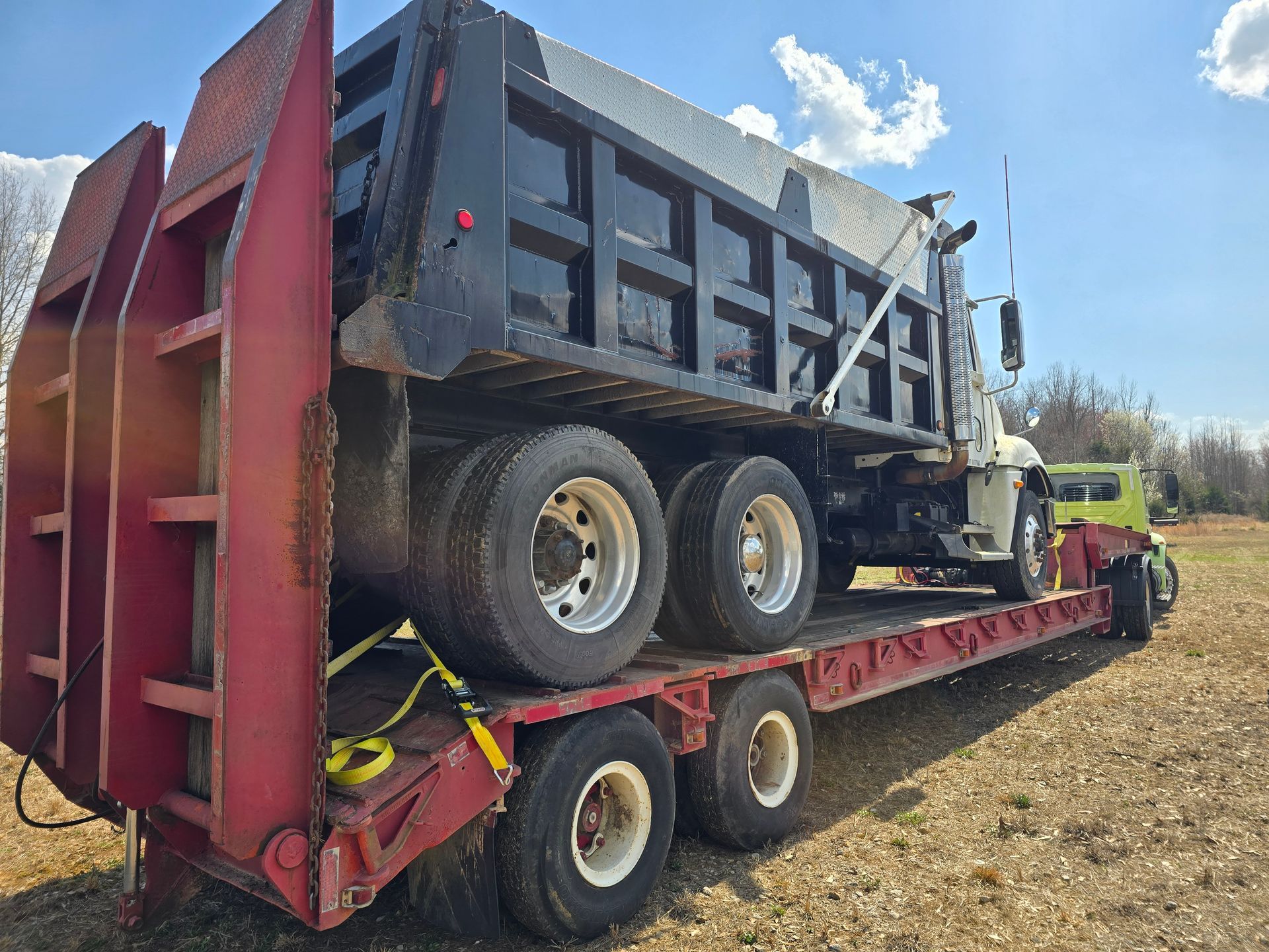 Red flatbed tow truck hauling a damaged black dump truck on a dirt lot under blue sky.