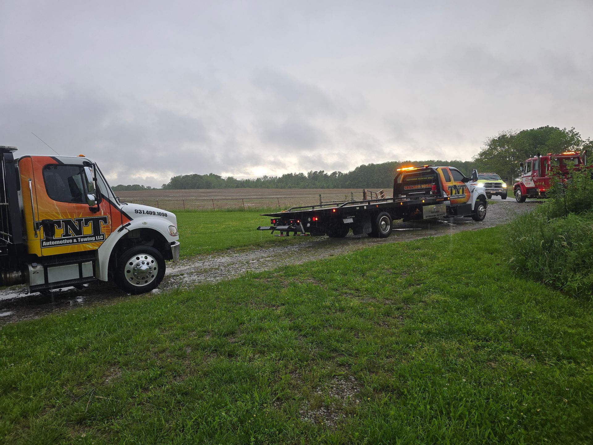 Tow truck and recovery vehicles on a muddy field road under cloudy skies.