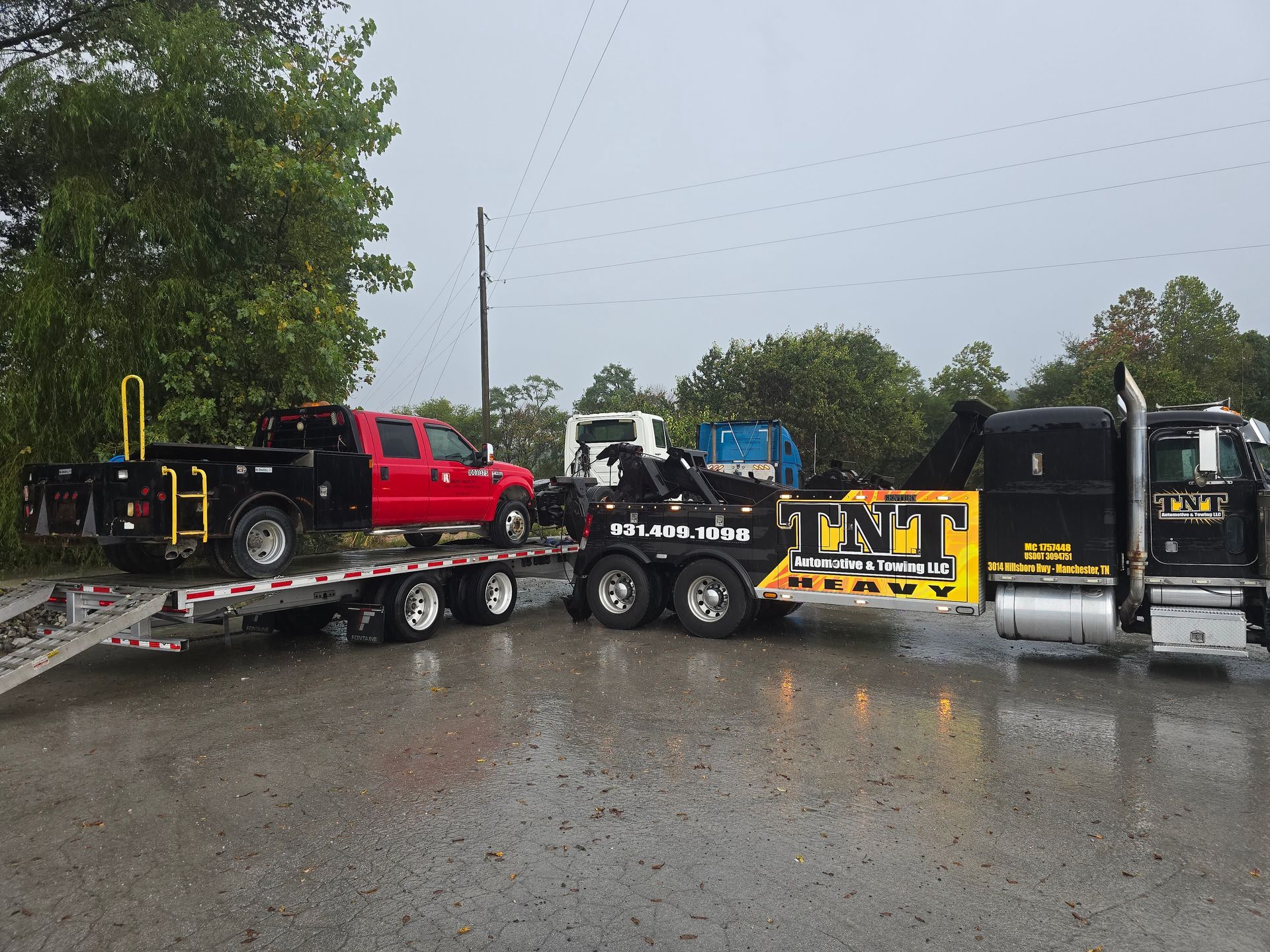 Tow truck hauling a red pickup and a yellow flatbed truck on a rainy roadside.