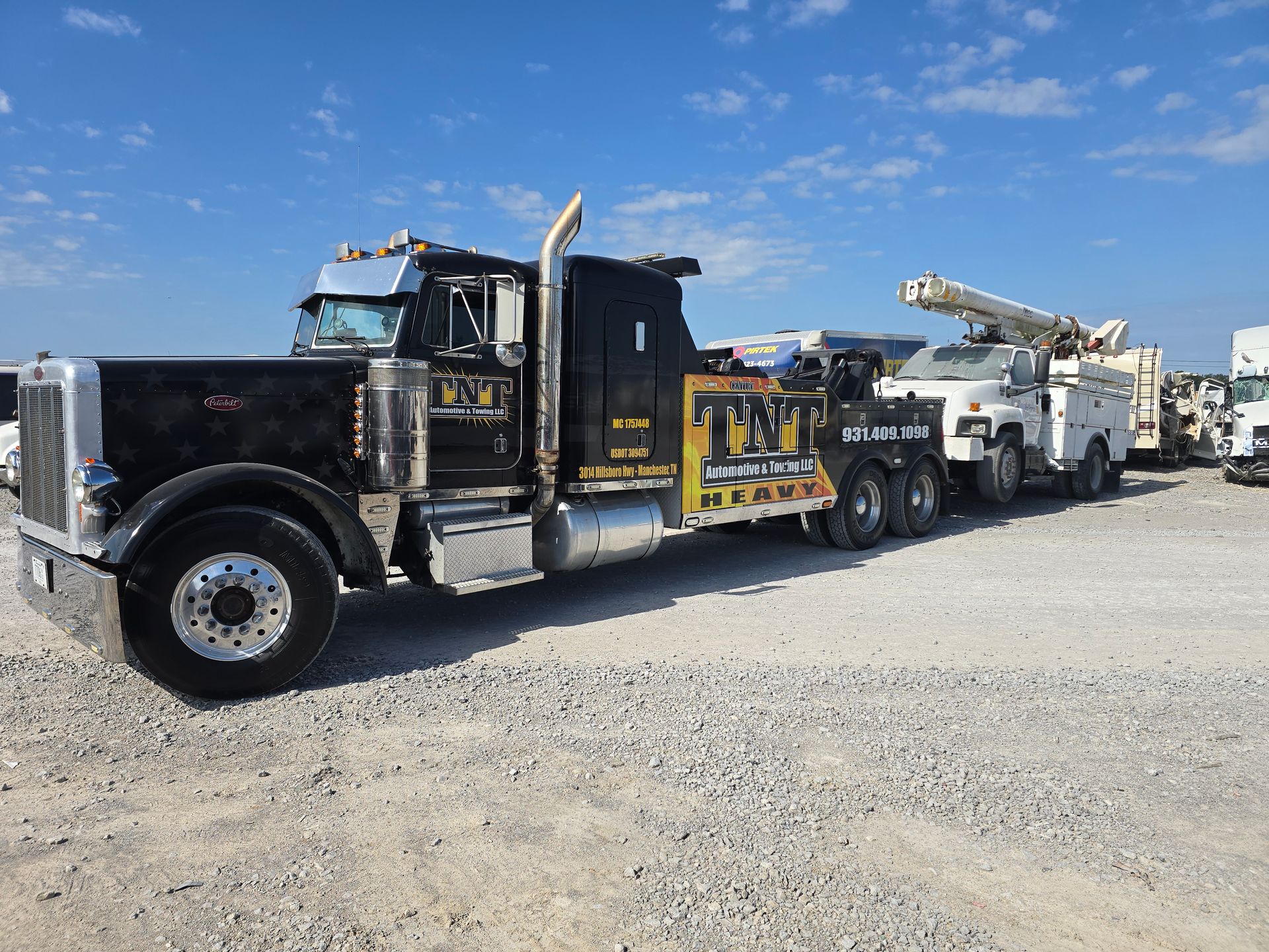 Black tow truck with equipment trailer parked on gravel under a blue sky.