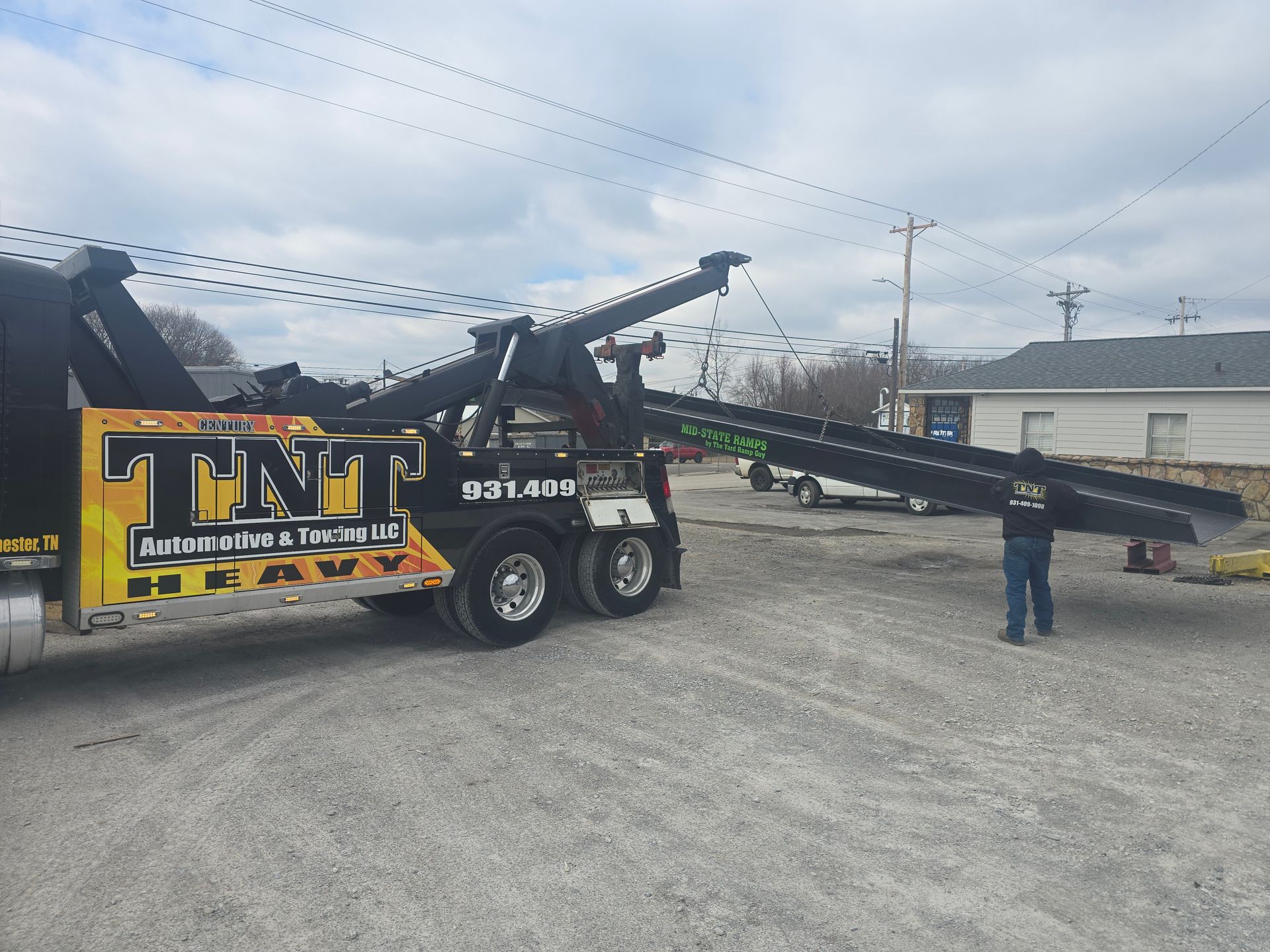 Tow truck with extended boom beside a yellow sign on a gravel lot, with a person standing nearby.
