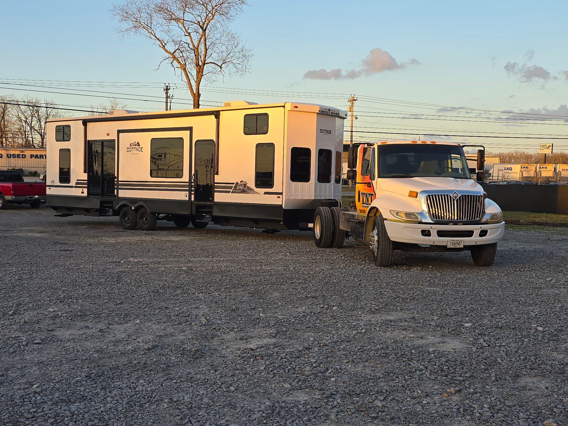 White and orange RV trailer hitched to a truck on a gravel lot at sunset.