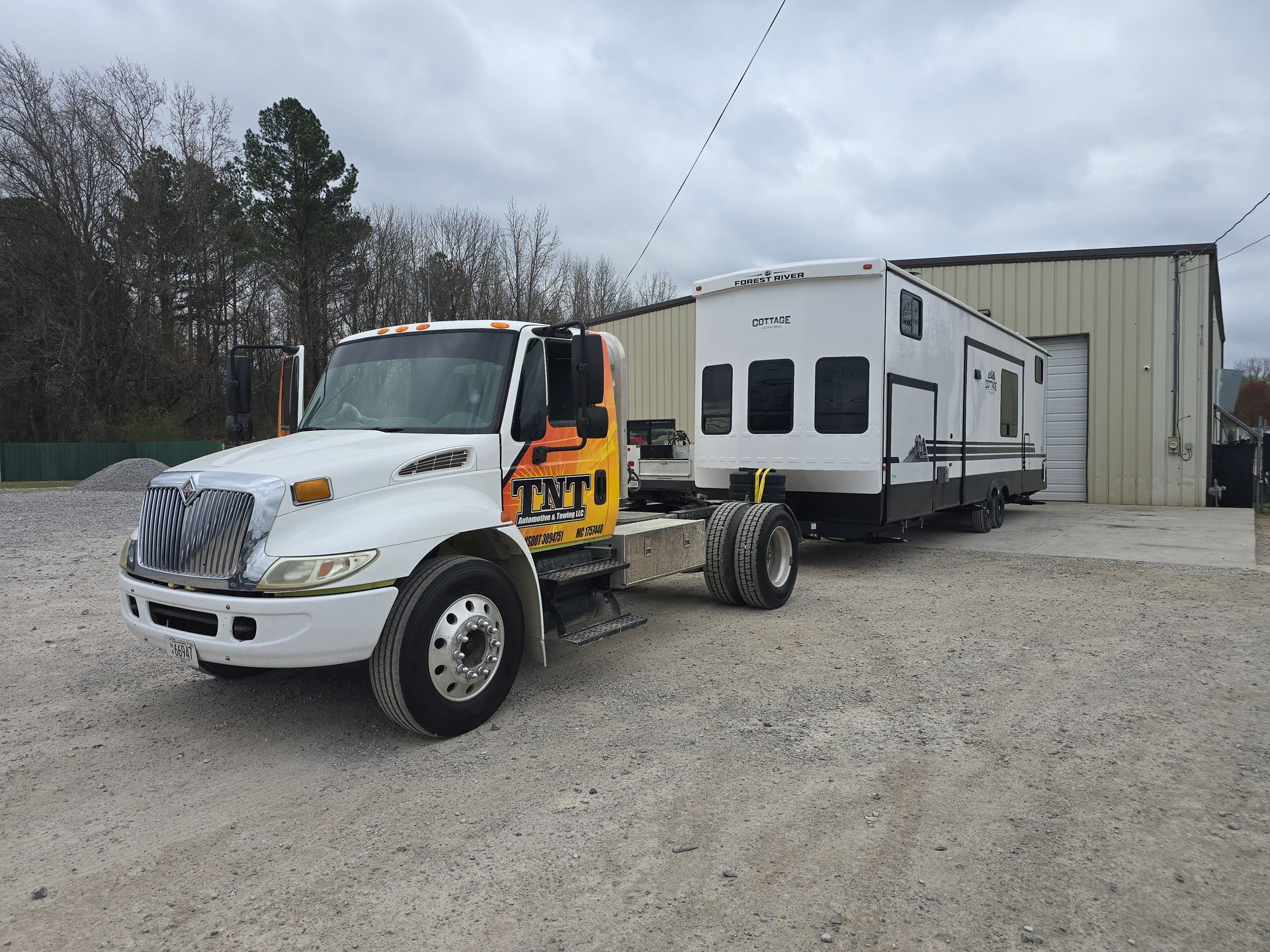 White tow truck hauling a white RV on a gravel lot beside a large shed.