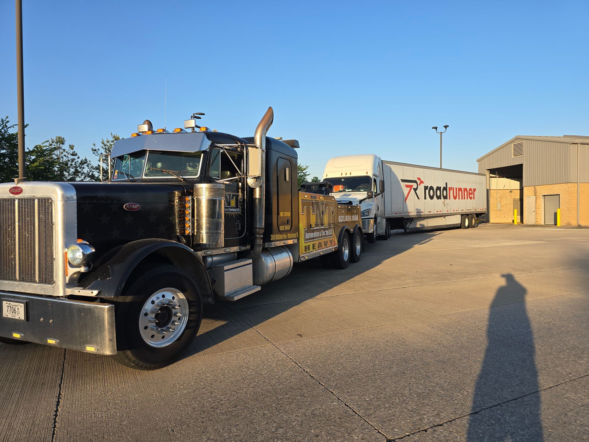 Black semi-truck hauling a white trailer in a sunny industrial lot under a clear blue sky.