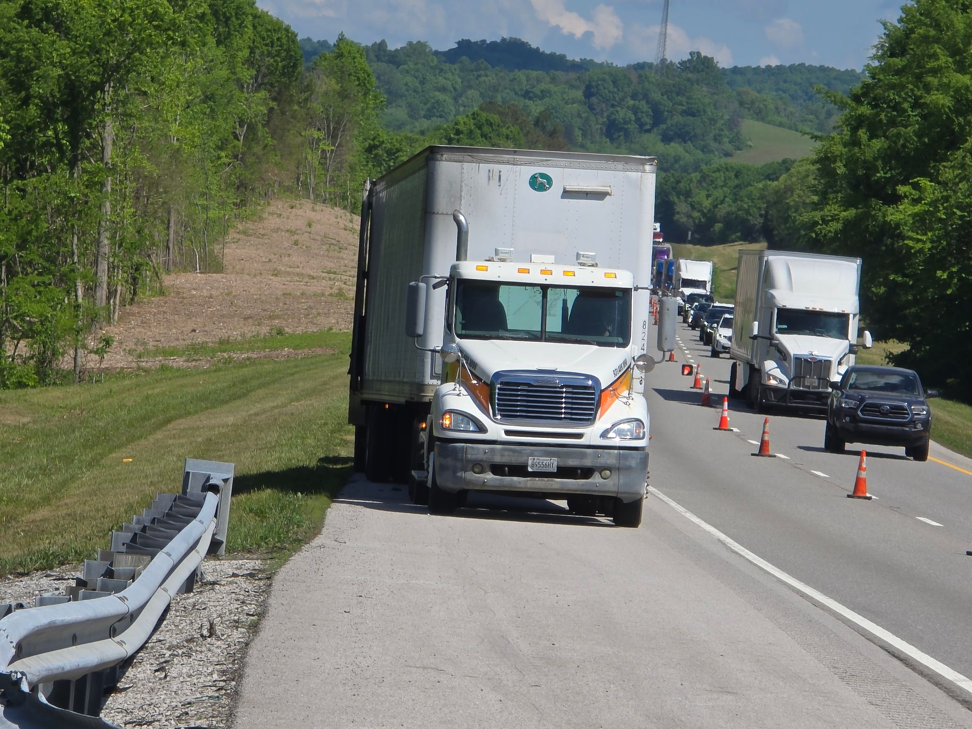 Disabled semi truck stopped on highway shoulder near accident cones, with cars and another truck in adjacent lane.
