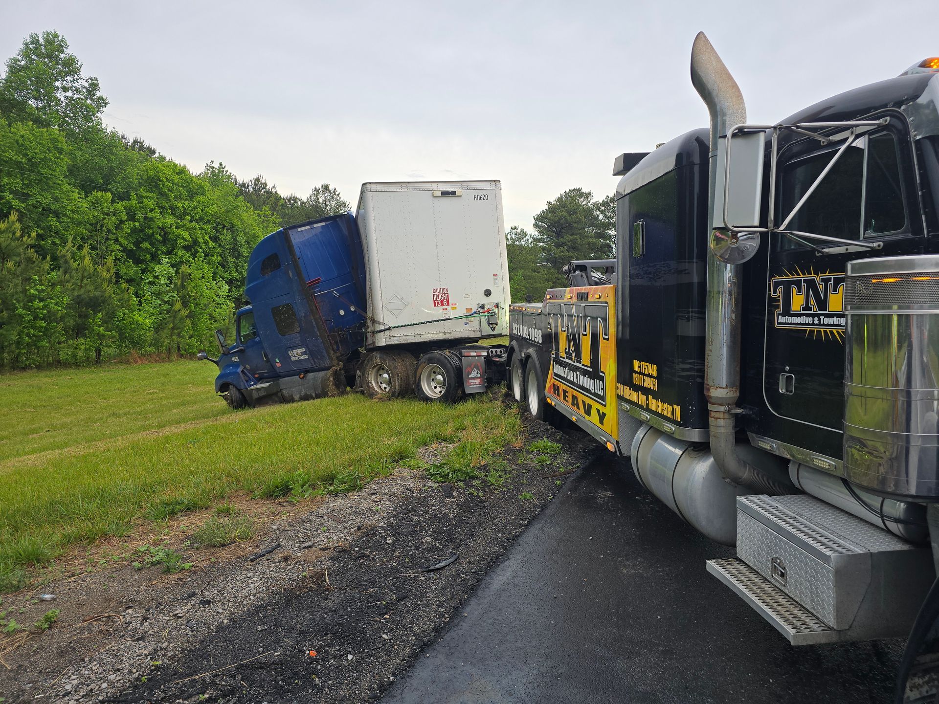 Two semi trucks pulled over on a wet roadside, one blue and one black with a white trailer.