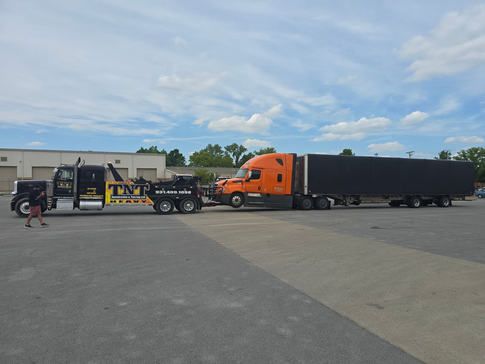 Orange semi truck with black trailer parked in a lot beside a warehouse, with people near a forklift.