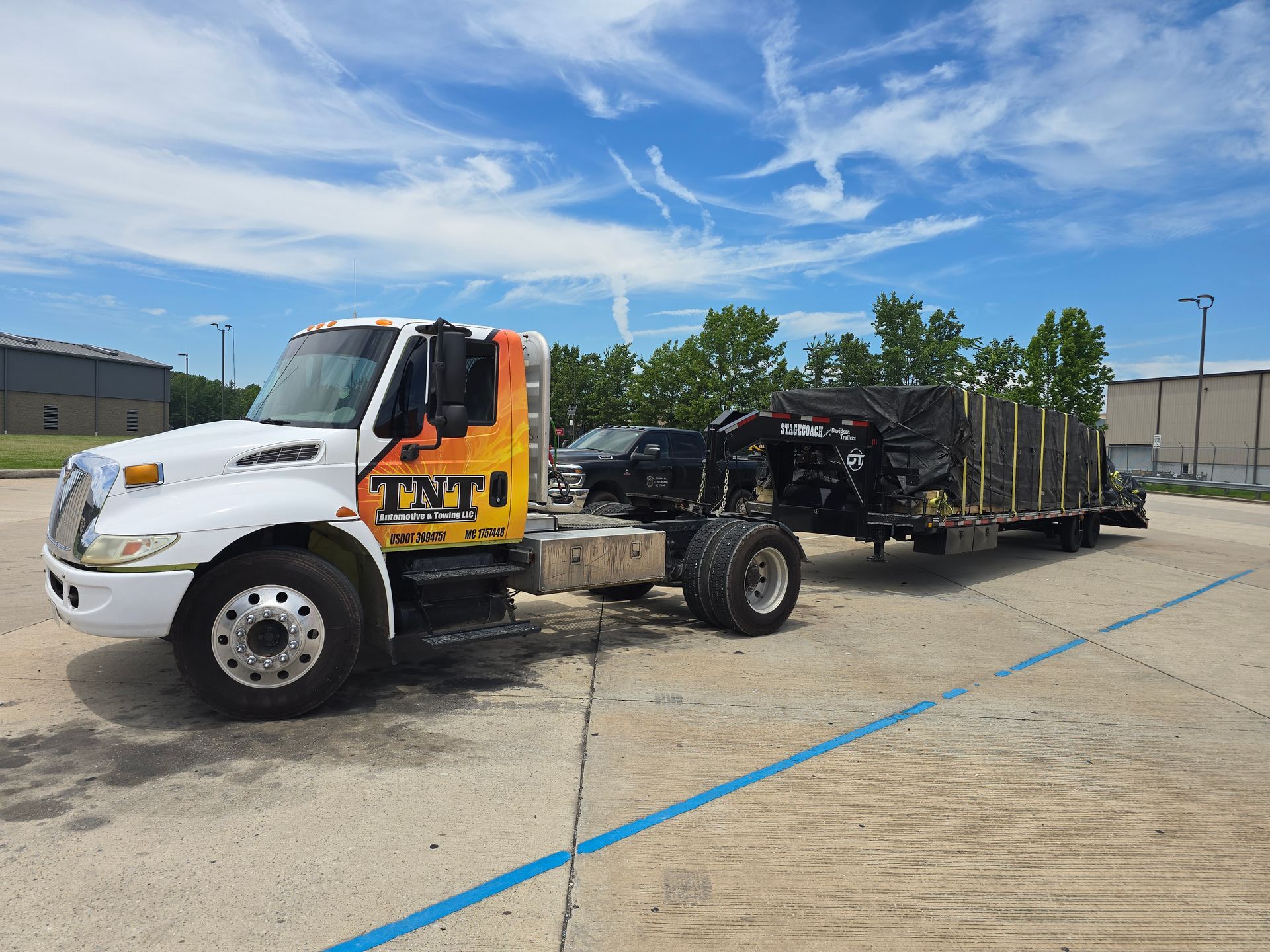 White tow truck hauling a black trailer in a sunny parking lot with trees and blue sky.