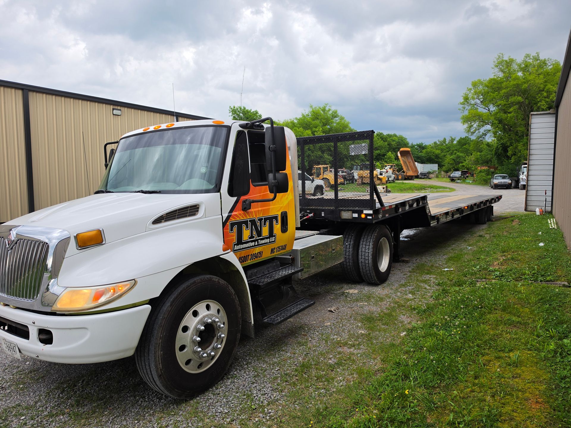 White flatbed tow truck parked on gravel beside a beige building under cloudy skies.