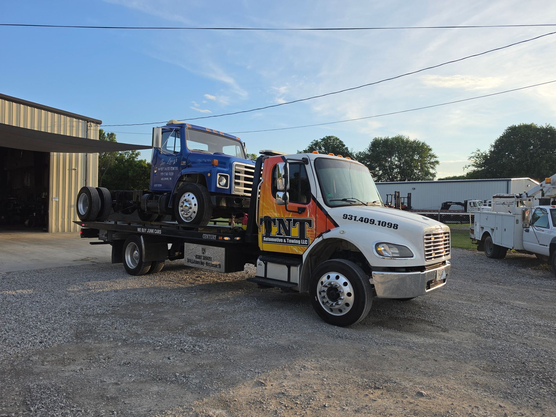 Tow truck hauling a blue off-road vehicle in a gravel lot near industrial buildings.