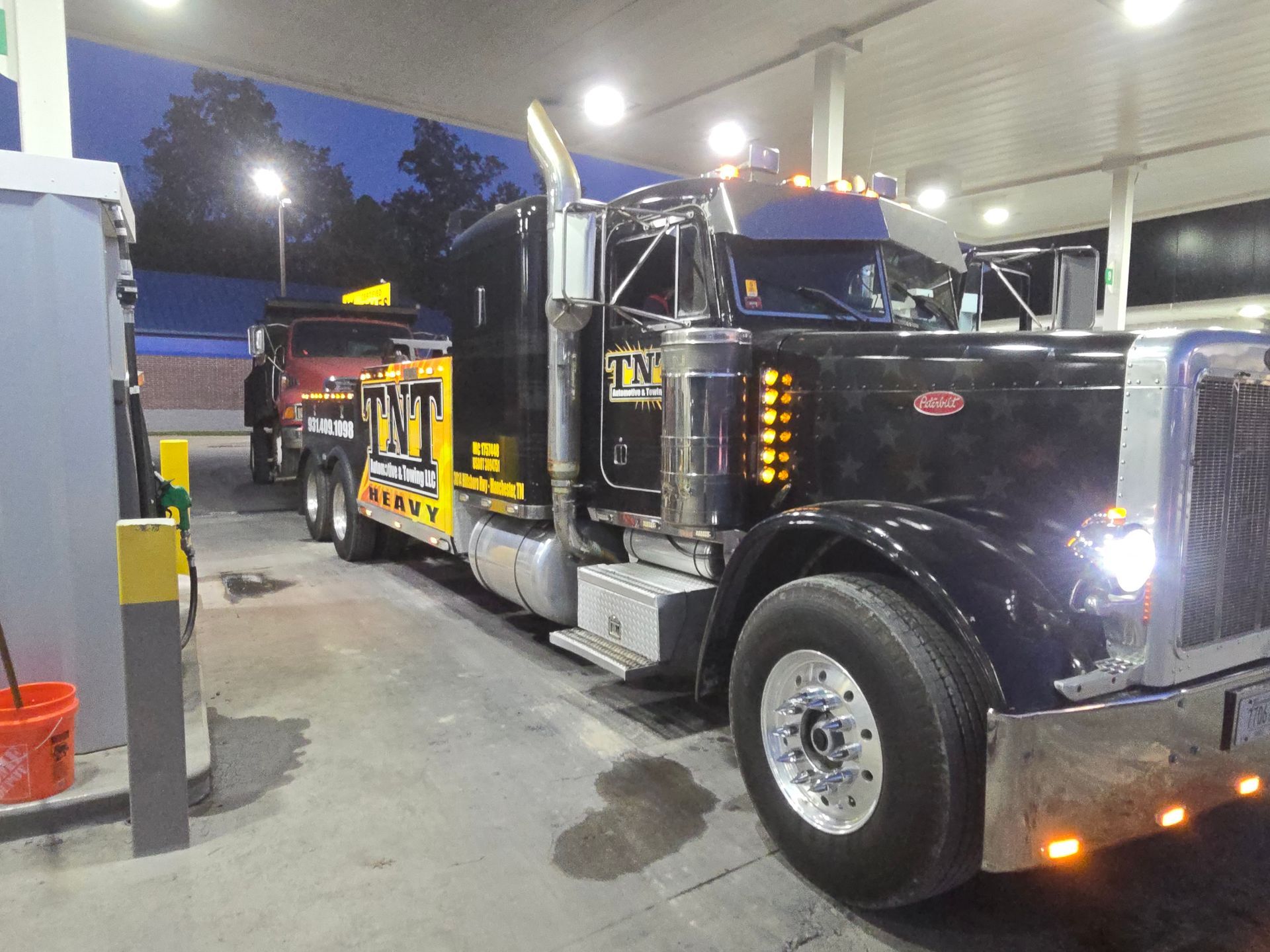 Black tow truck at a gas station pump at night, with another truck behind it.