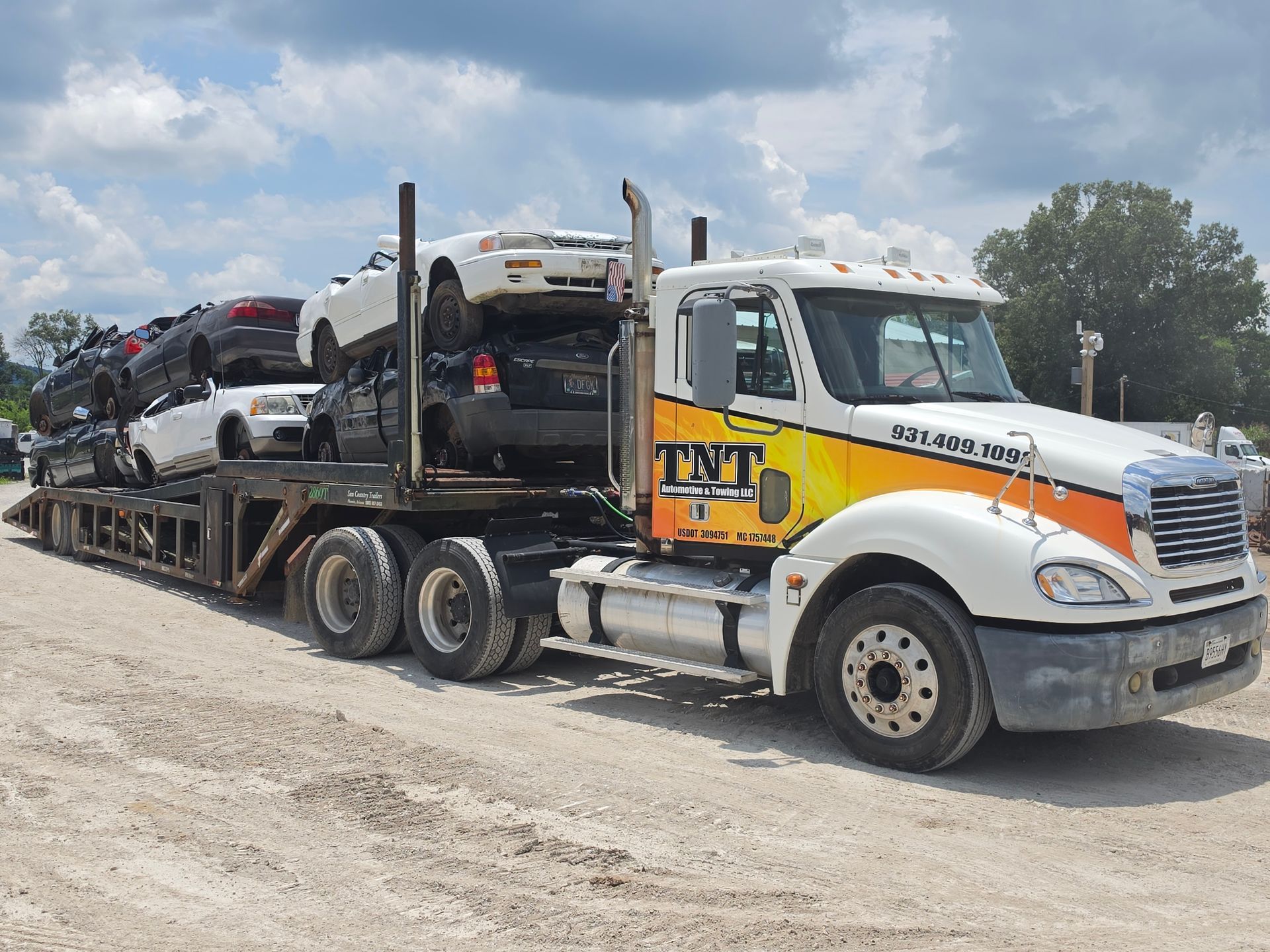Tow truck hauling a damaged car on a flatbed in a gravel lot under a cloudy sky.