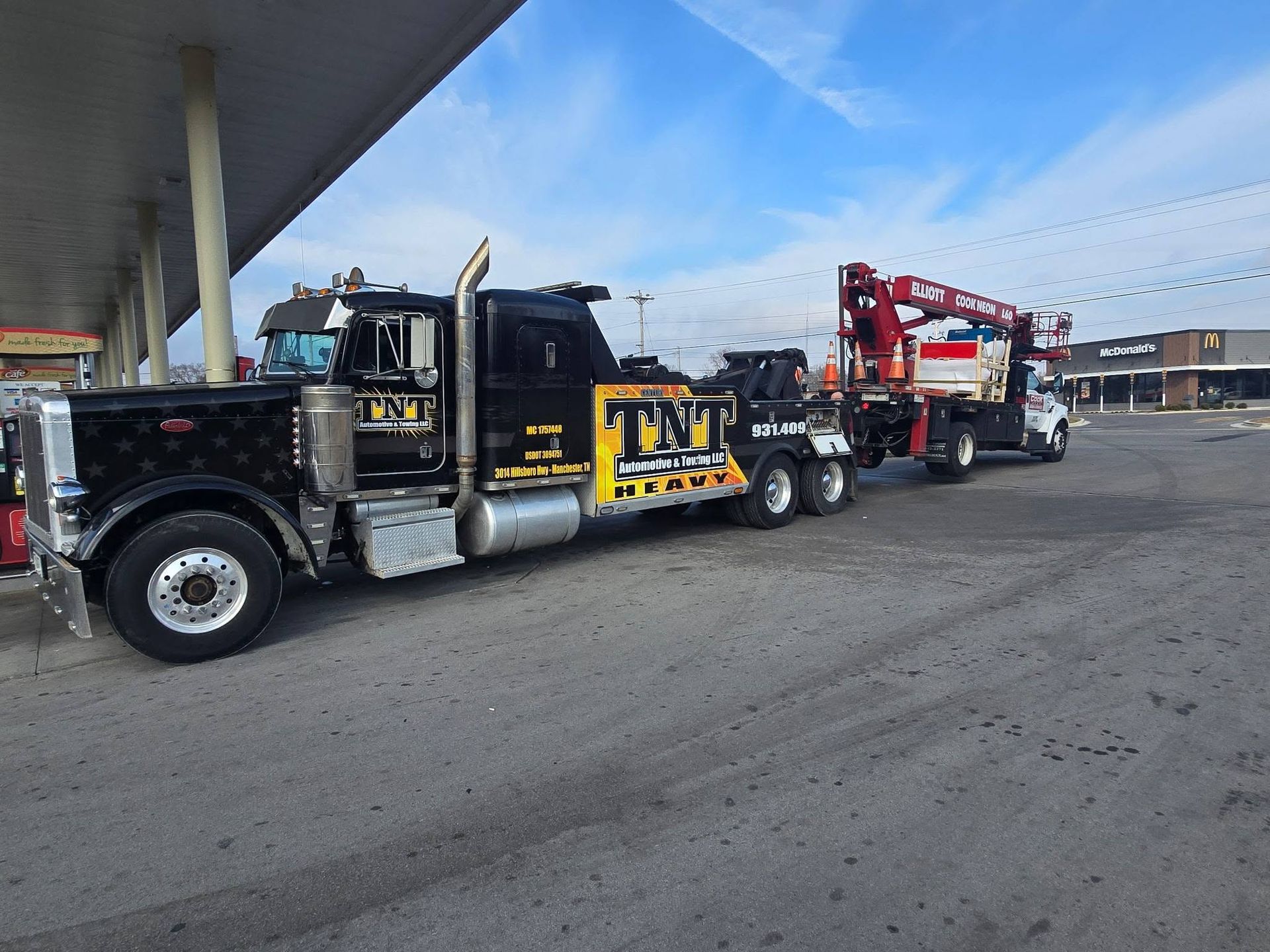 Black semi-truck hauling a red crane trailer in an industrial lot under a highway overpass.