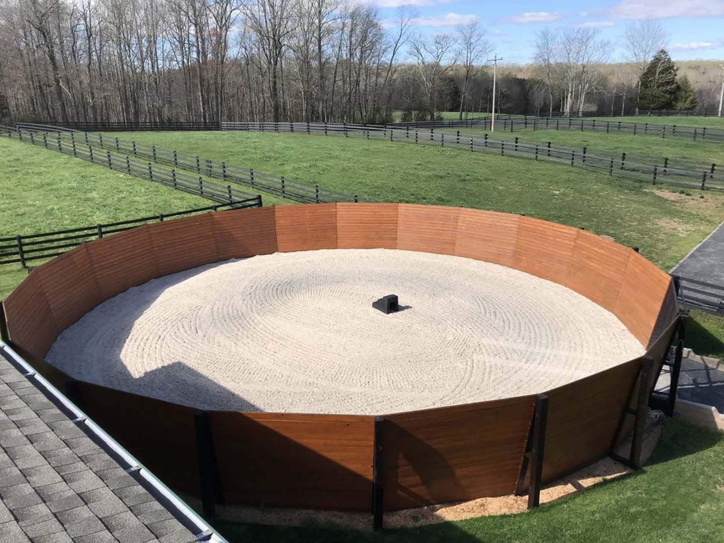 Circular riding arena with tan sand, surrounded by a brown wooden fence, in a grassy field.