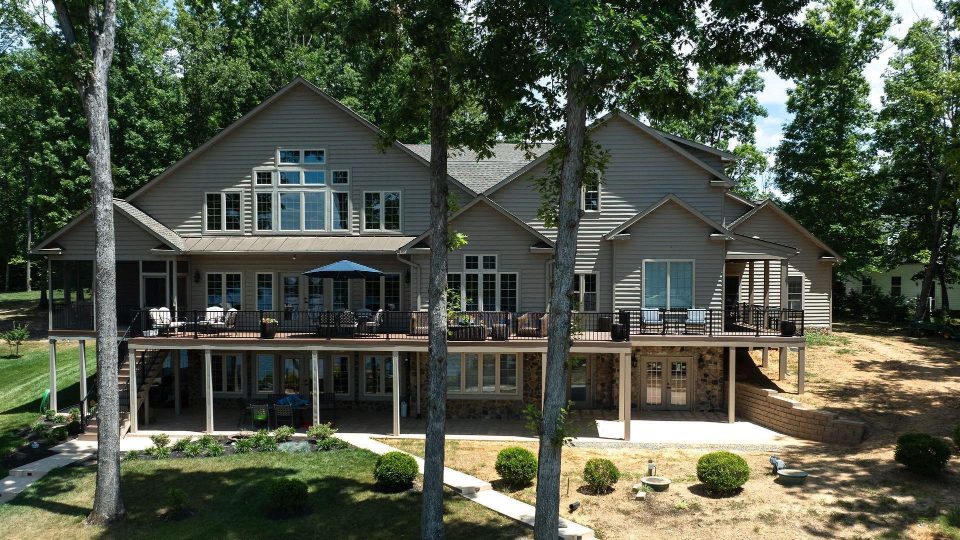 Large house with a wrap-around deck on a hill overlooking a tree-lined area.