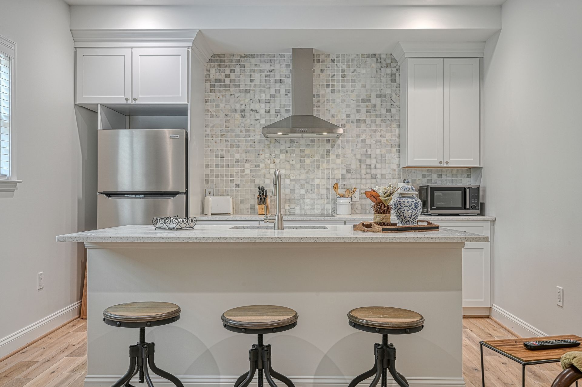 Kitchen with a white countertop island and three stools. Stainless steel appliances and gray backsplash.
