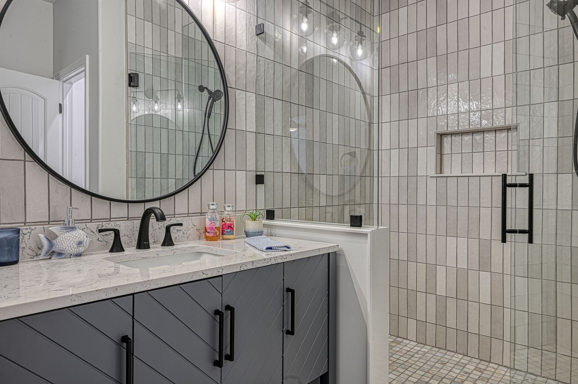 Bathroom with gray vanity, round mirror, and shower with light-colored, vertical tile.