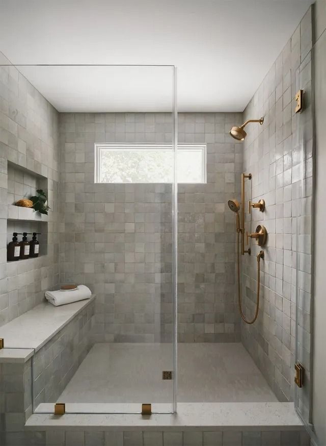Modern shower with gray tiled walls, glass door, bench, and gold fixtures.