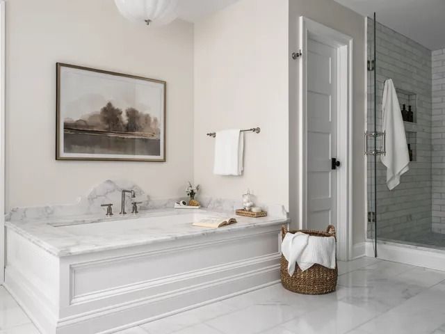 Elegant bathroom with marble tub, art, and glass shower. White walls, wood basket.