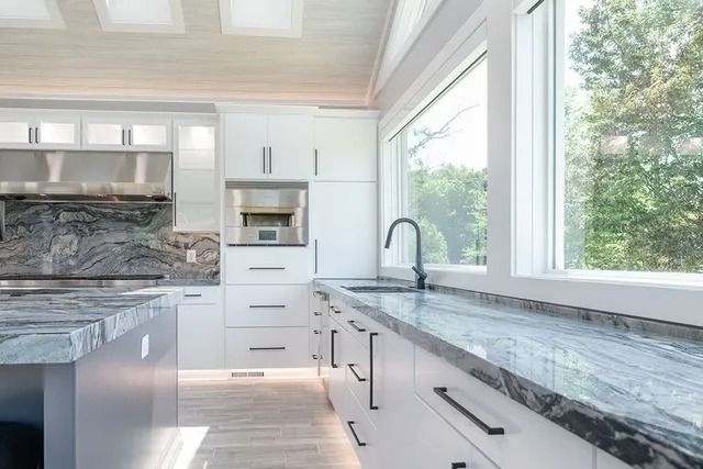 Modern white kitchen with marble countertops, large windows, and dark fixtures.