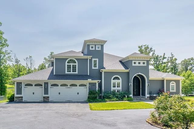 Gray stucco house with three-car garage, arched windows, and a dark roof under a blue sky.
