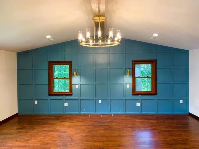 Empty room with blue paneled accent wall, two brown-framed windows, and gold chandelier.