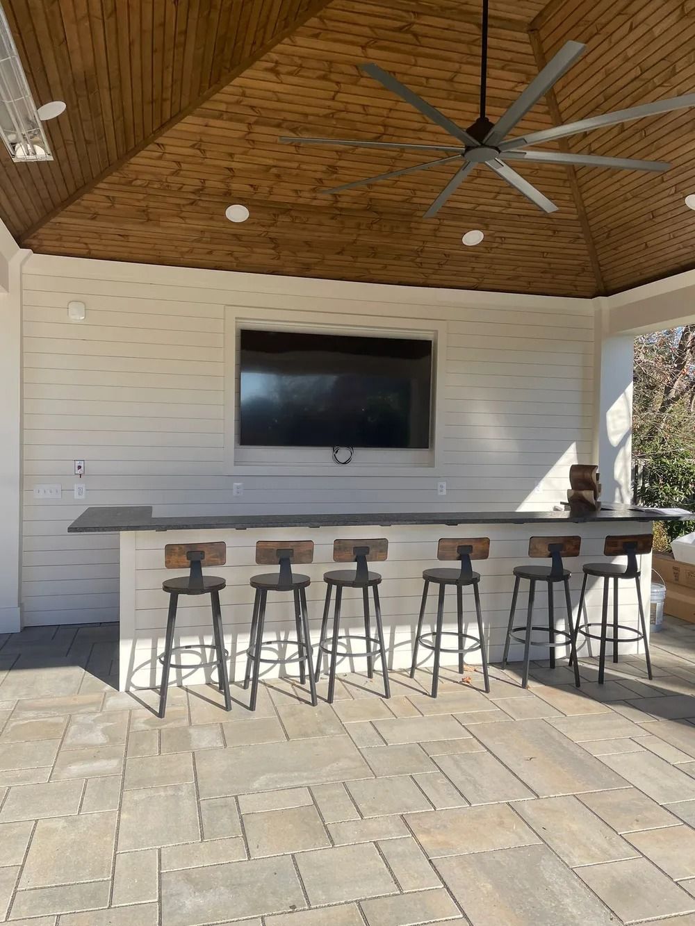 Outdoor bar with a TV, stools, and ceiling fan. Light wood ceiling, white walls, and stone patio.