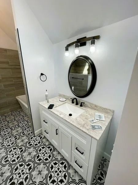Bathroom with white vanity, black hardware, patterned tile, round mirror, and wood-look light fixture.