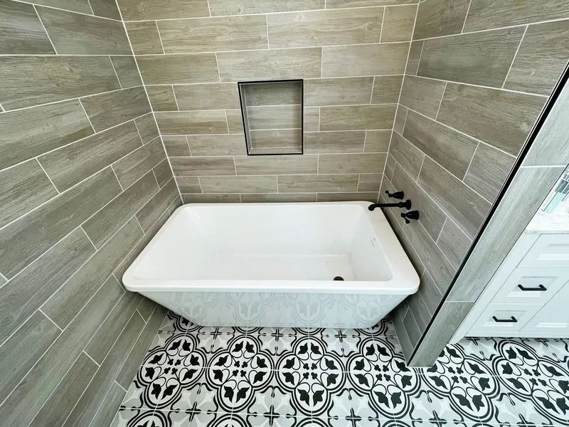 Bathroom with a white tub, gray tiled walls, decorative floor tiles, and black fixtures.