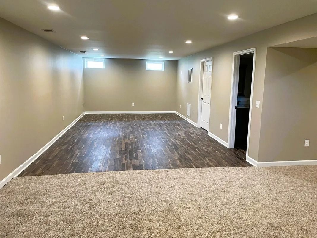 Empty basement room with wood-look flooring and beige carpet, light walls, recessed lighting, and a doorway.