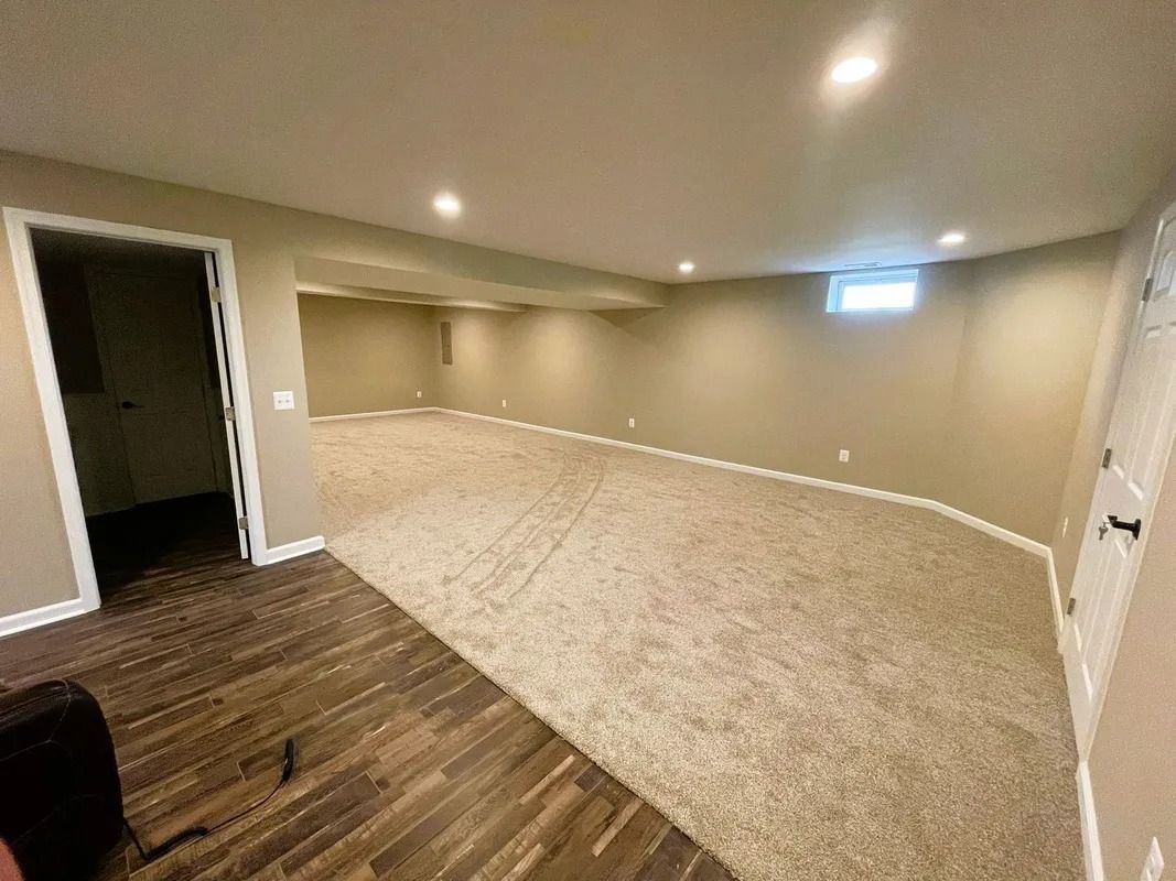 Empty basement room with tan walls, carpet, and a small window. There are two doorways.