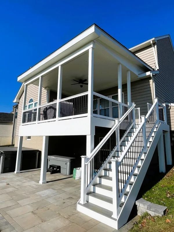 White screened porch with stairs attached to a beige house, overlooking a stone patio, with black railings.