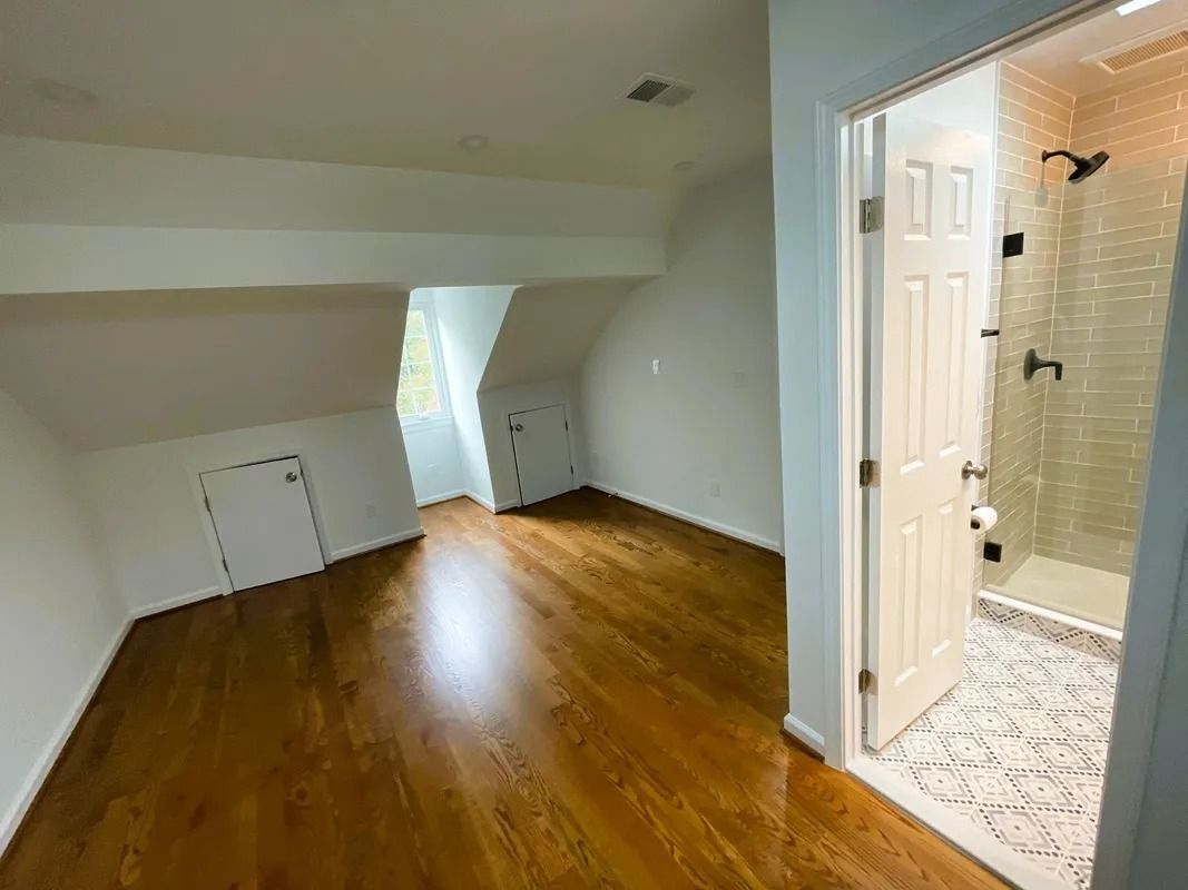 A room with hardwood floors, a sloped ceiling, and an open bathroom door; white walls.