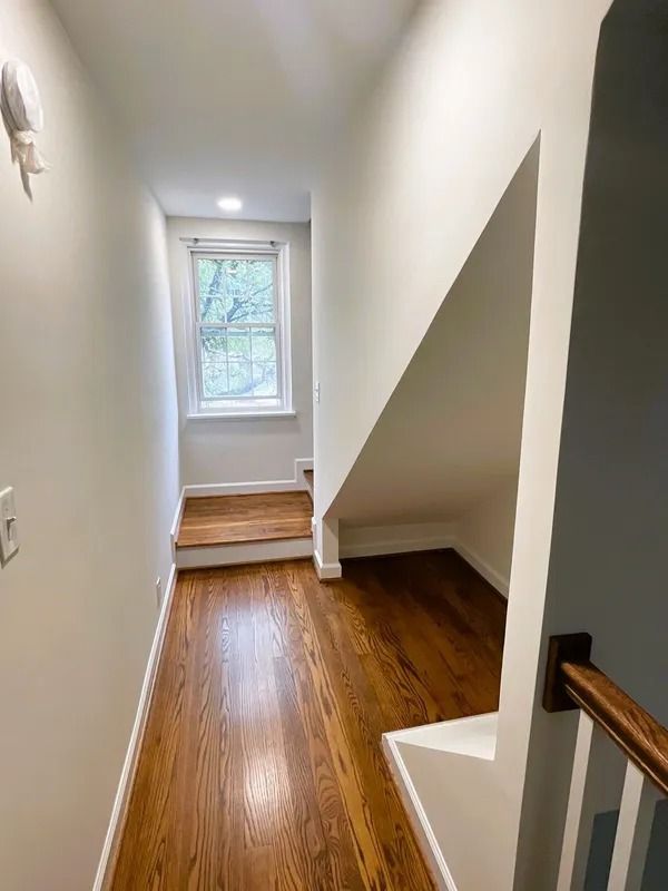 Narrow hallway with hardwood floor, angled wall, window, and small staircase.