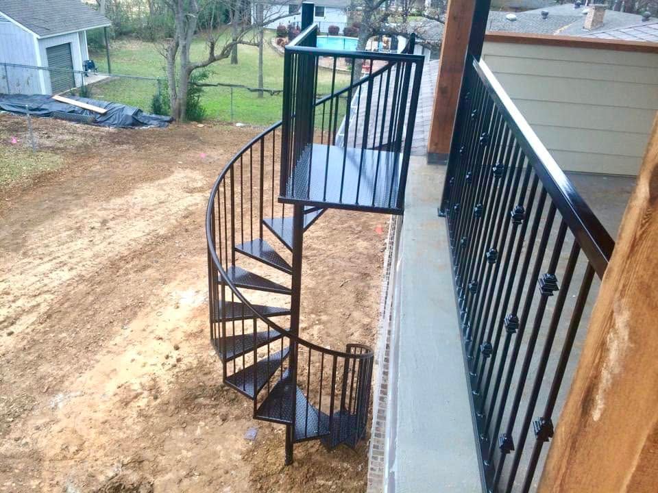 Black metal spiral staircase beside a house on a dirt slope, leading to an elevated deck.