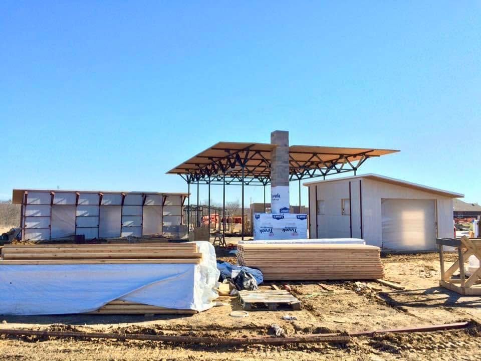 Small industrial site with stacked panels, a roofed structure, and a white building in a dusty lot under a blue sky.