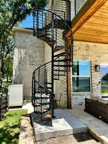 Black metal spiral staircase beside a stone house and wooden deck in a sunny yard.