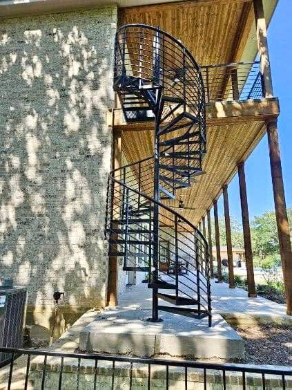 Black spiral staircase beside a building, leading to a raised walkway in a sunny outdoor area.