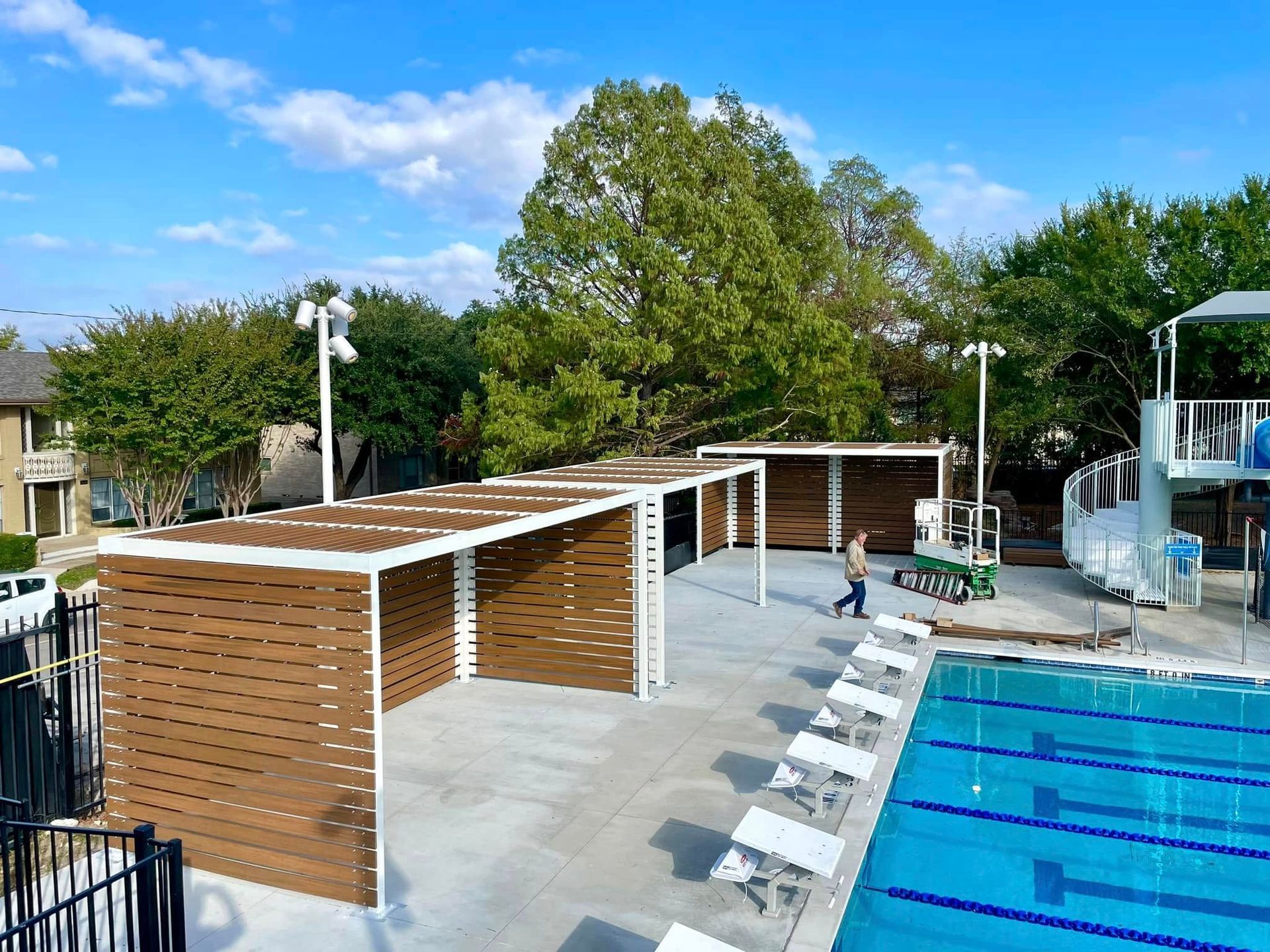 Outdoor pool area with covered lounge cabanas, white chairs, and blue water under a bright sky.