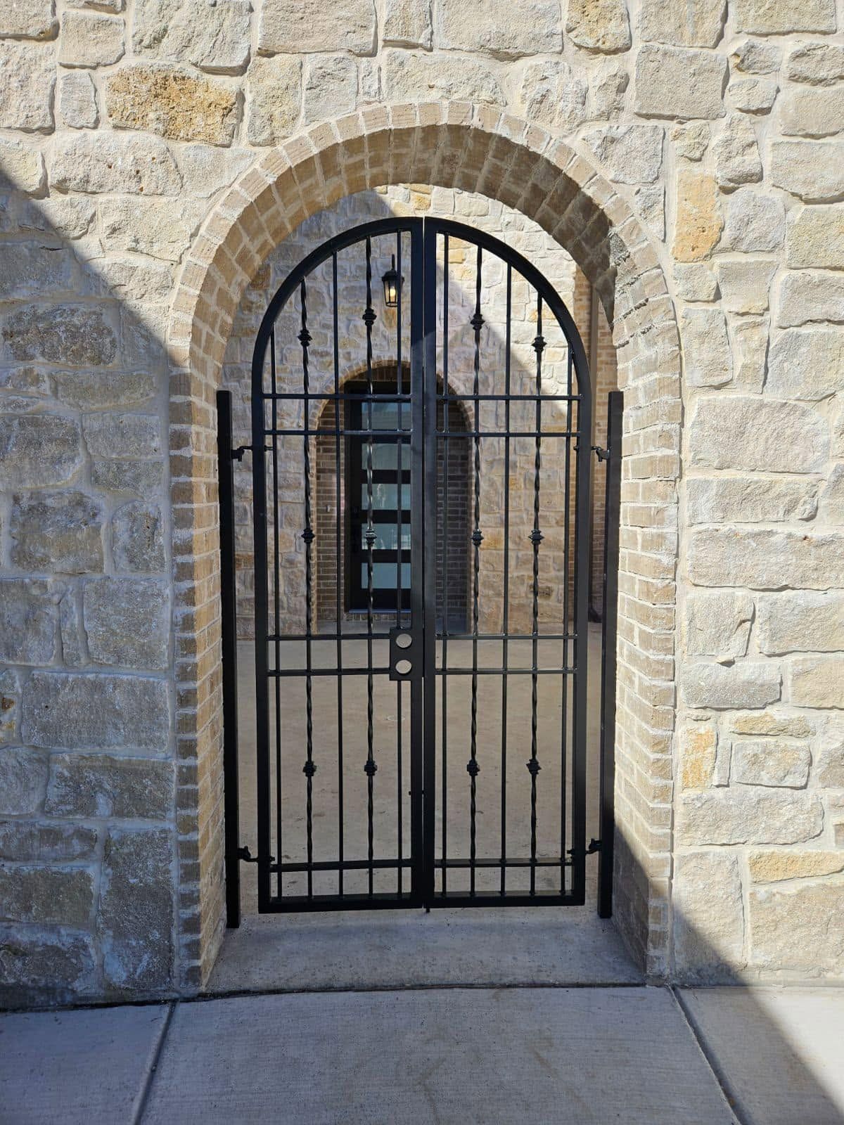 Black iron gate set in a stone archway and wall, with a narrow doorway behind it.