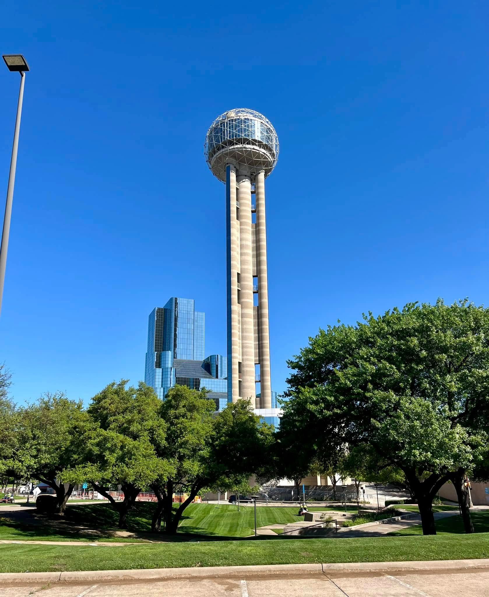 Park with green trees in foreground and a tall tower with a spherical top against a blue sky.