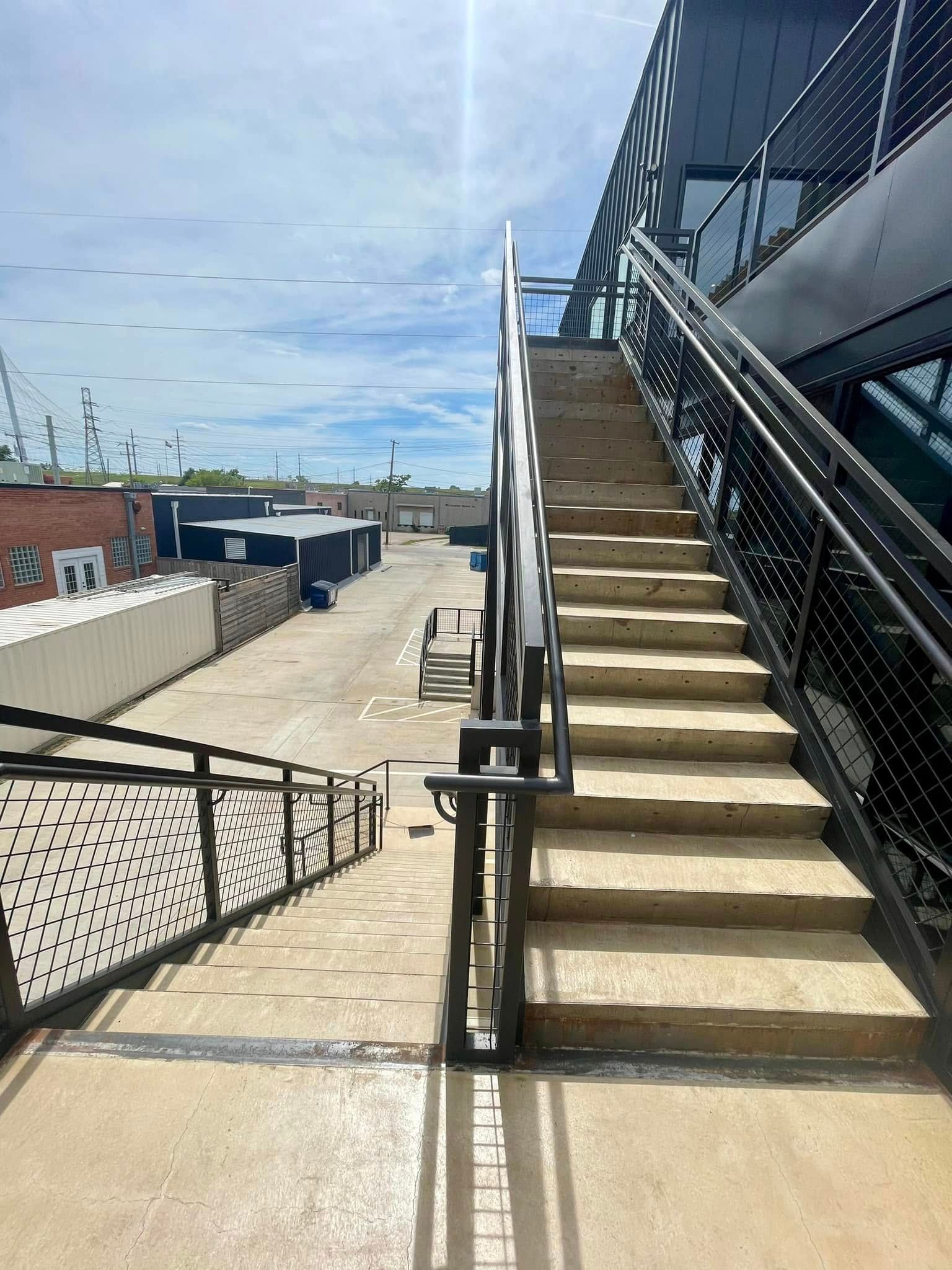Outdoor concrete stairway with black railings beside a rooftop walkway under a partly cloudy sky.