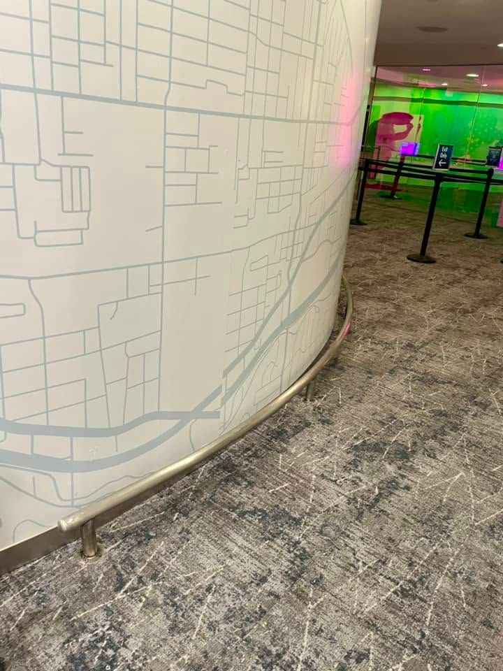 Curved white wall with pale map-like lines beside a carpeted walkway and neon-lit area beyond barriers.