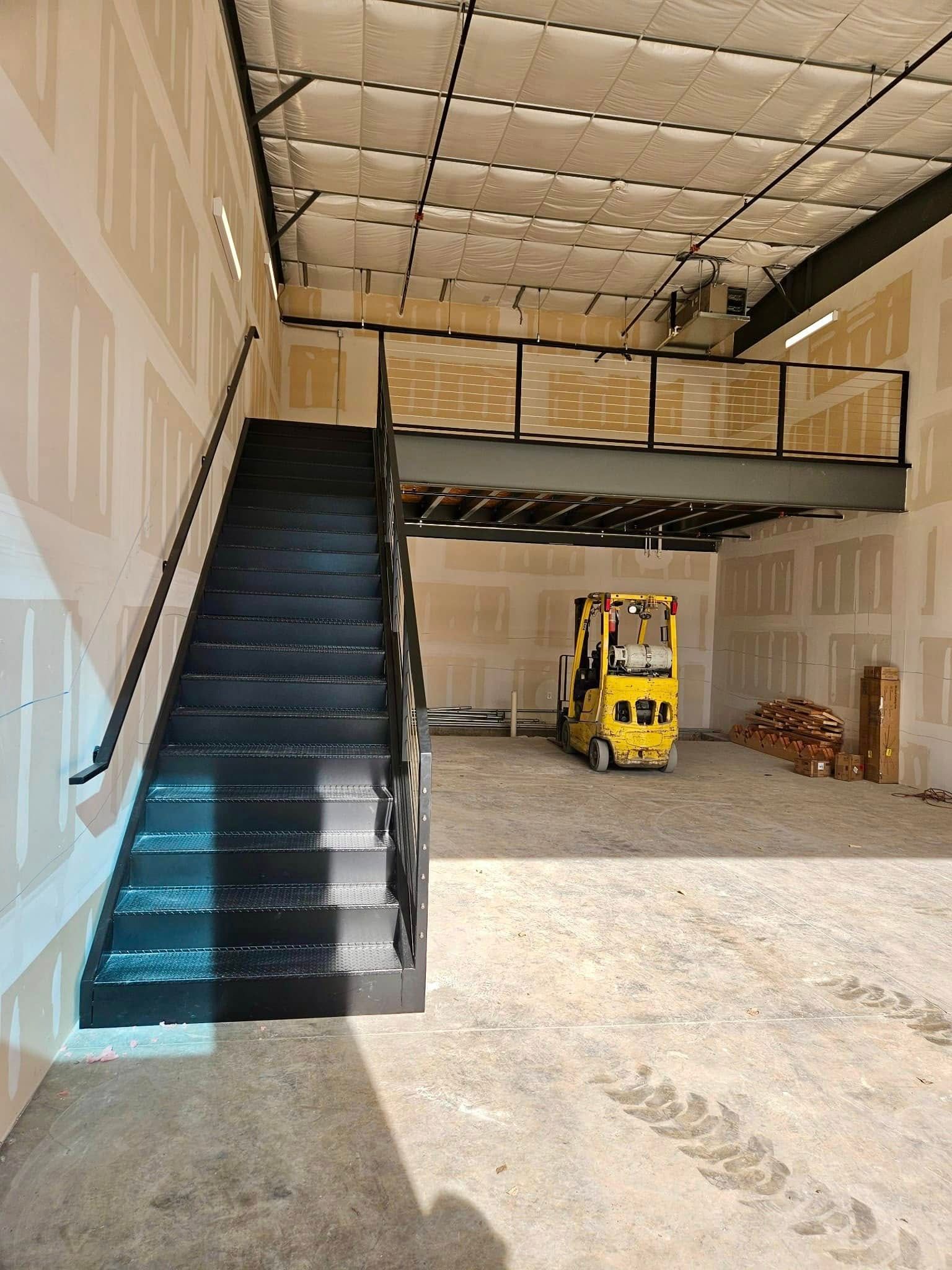 Interior of an unfinished warehouse with a metal staircase, mezzanine, and a yellow forklift on a concrete floor.
