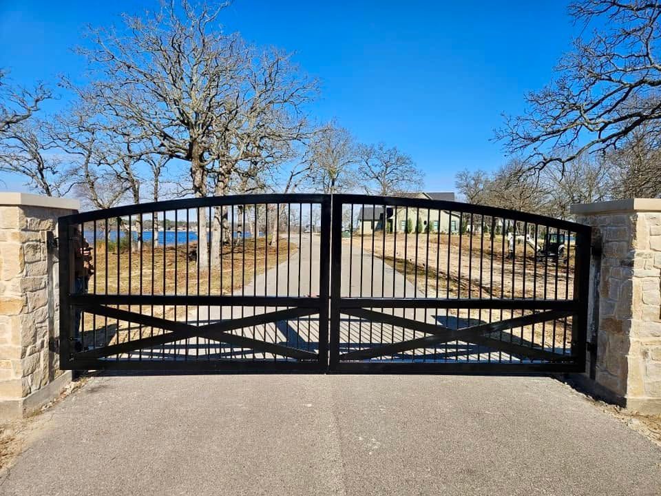 Black metal double gate between stone pillars across a driveway under a clear blue sky.
