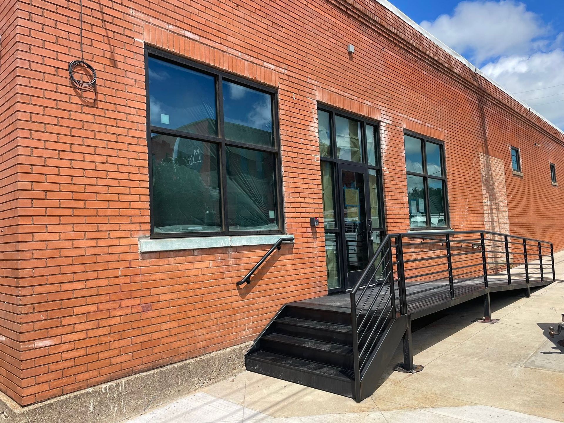 Brick building exterior with black-railed ramp, steps, and large windows under a blue sky.