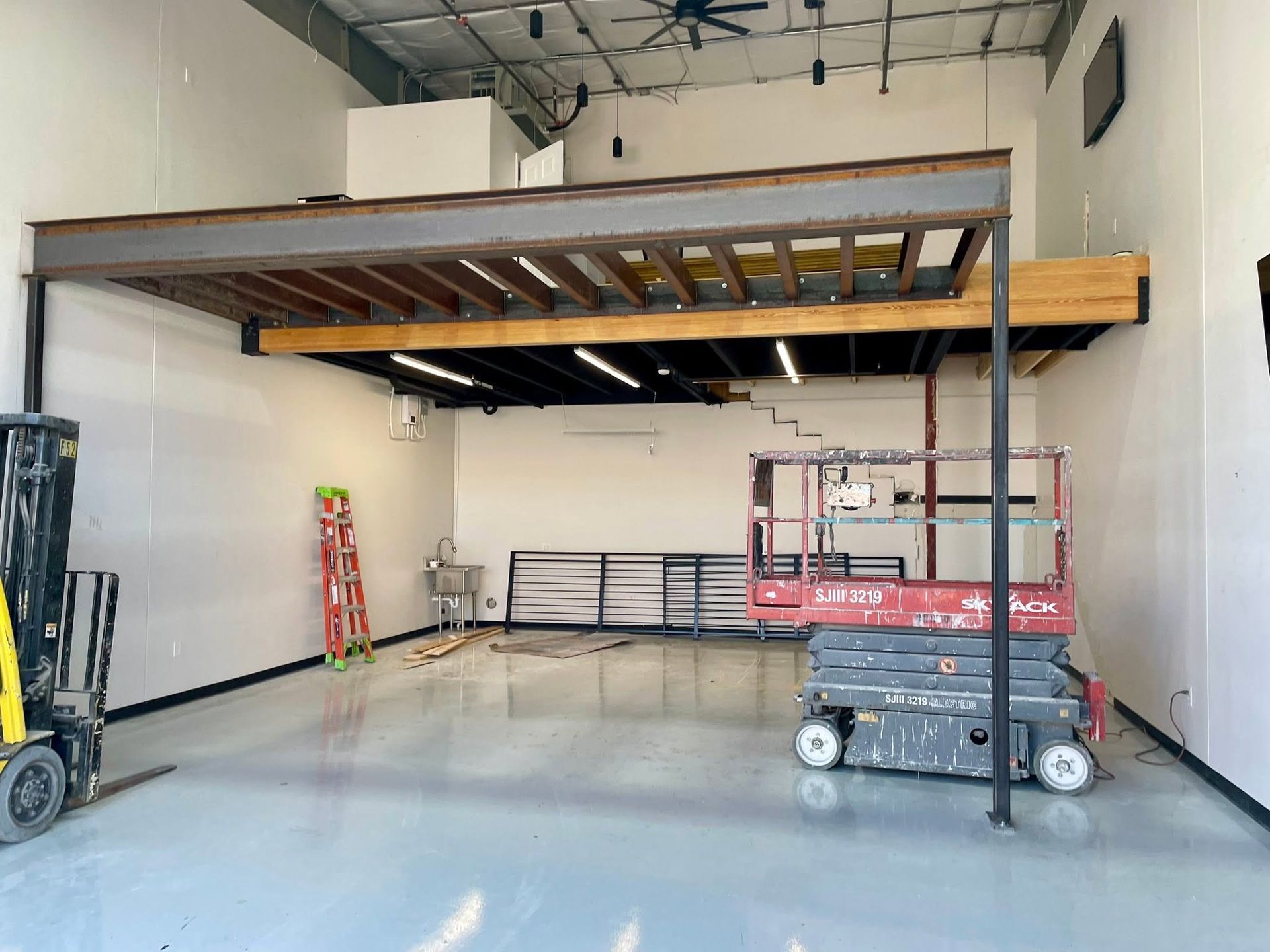 Empty industrial room with a raised loft/mezzanine, ladders, railings, and a lift on a polished concrete floor.