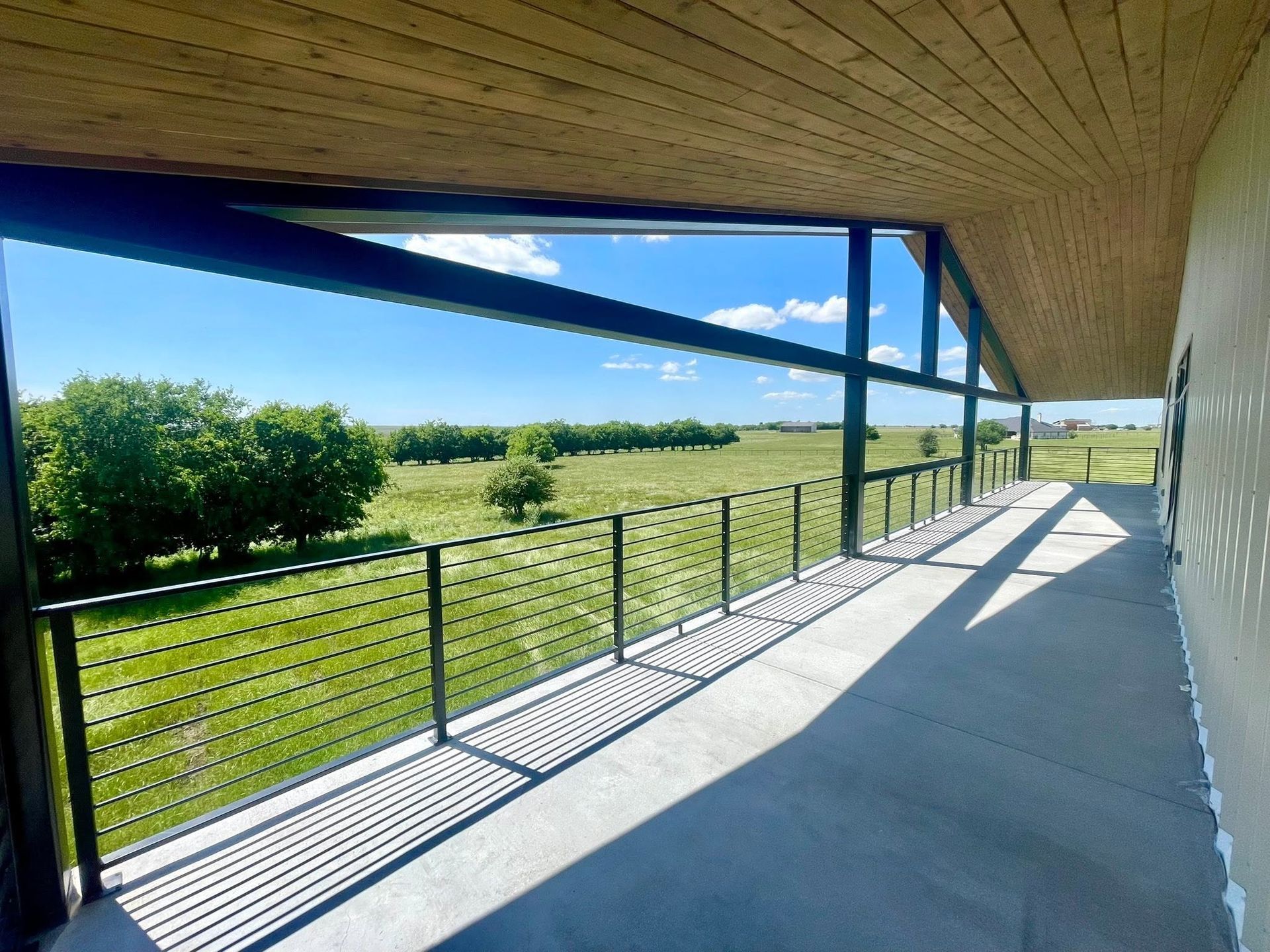 Covered patio overlooking green fields with black railing and long shadows in sunlight.